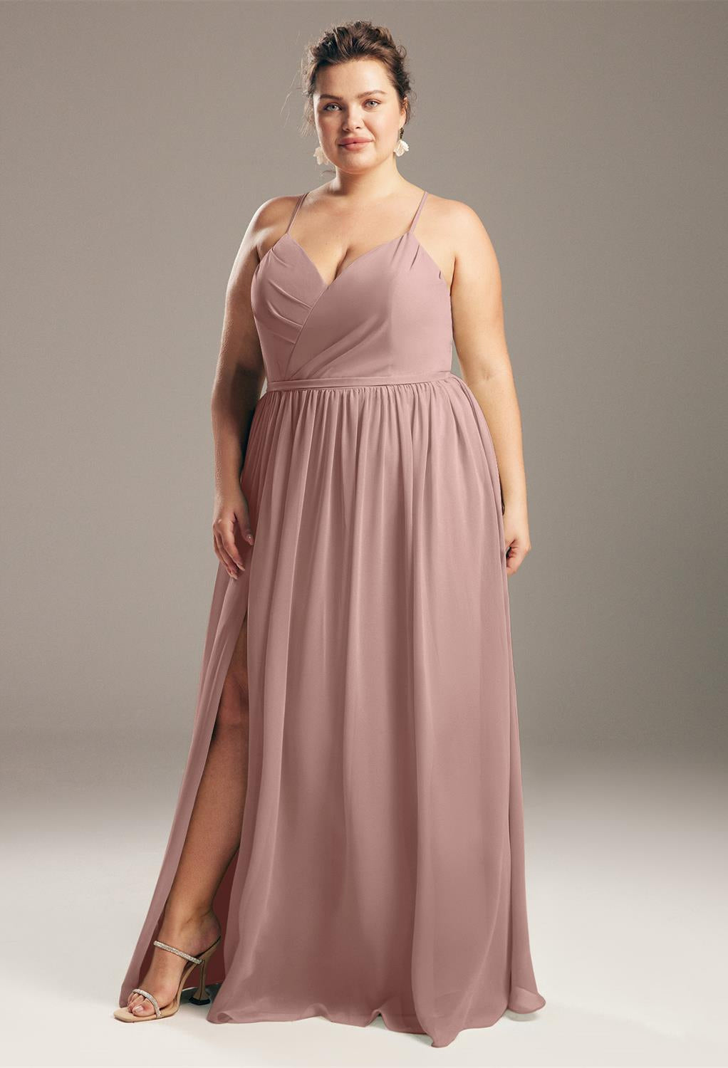 A woman, in the budget-friendly Wilfreda chiffon bridesmaid dress from Bergamot Bridal, featuring a mauve hue, a high slit, and spaghetti straps, stands gracefully against a plain gray backdrop, echoing the classic sophistication of bridal boutiques in London, Ontario.