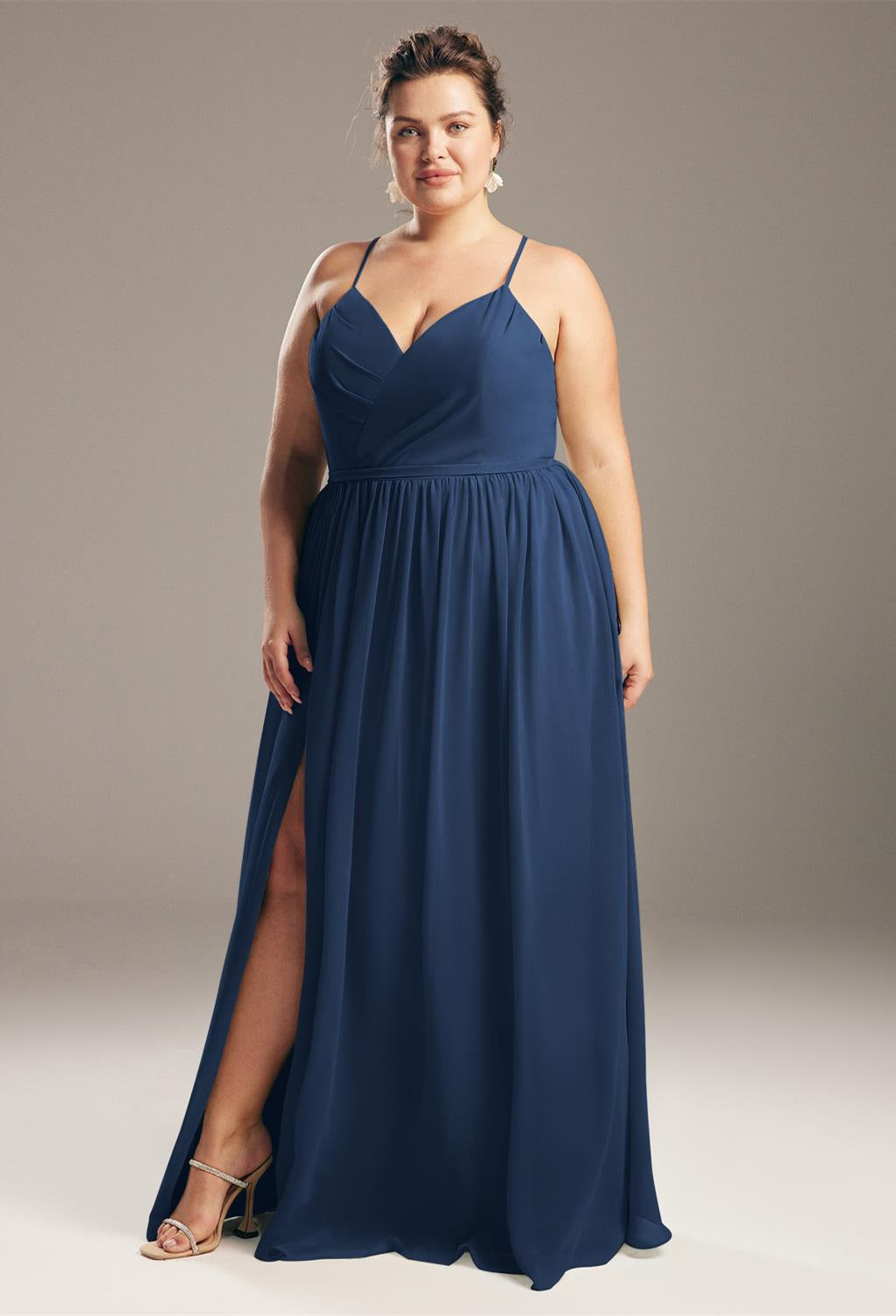 A woman in a blue Wilfreda Chiffon Bridesmaid Dress - Off The Rack, standing confidently against a gray background, her hand resting on her hip, reminiscent of styles seen in Bergamot Bridal.