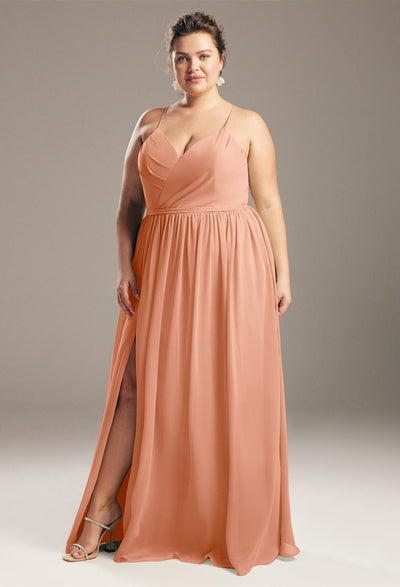 A person wearing the Wilfreda chiffon bridesmaid dress by Bergamot Bridal, featuring a peach color, sleeveless design, V-neckline, and high slit, stands gracefully against a neutral background.