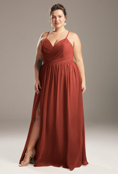 Against a gray backdrop, someone stands gracefully in a full-length, sleeveless dress in rust with a slit. This striking outfit would seamlessly enhance the range of bridesmaid dresses offered by Bergamot Bridal's Wilfreda collection at their premier bridal shop in London, Ontario.