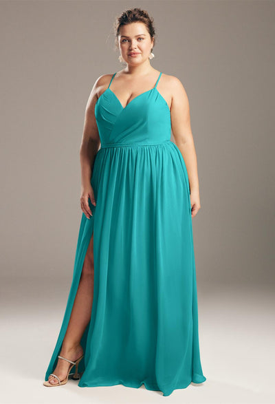 A woman in a teal Wilfreda - Chiffon Bridesmaid Dress - Off The Rack stands confidently, posing with one hand on her hip and a slight smile from Bergamot Bridal.