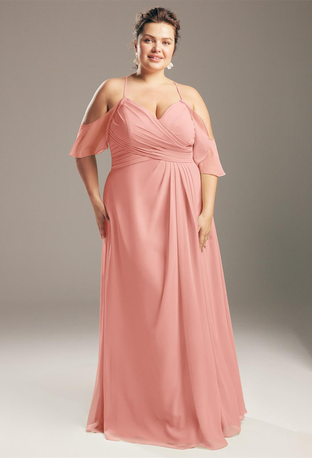 A woman in a pink Jenifer chiffon bridesmaid dress from Bergamot Bridal stands gracefully against a gray background, evoking the elegant style found at a bridal shop in London.