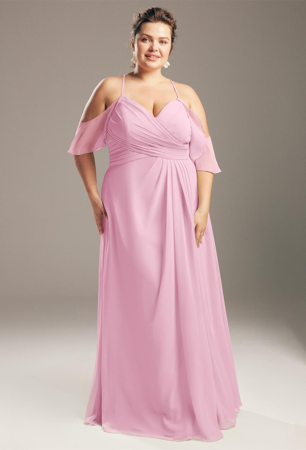 A person poses in a floor-length, pink gown with off-the-shoulder straps, reminiscent of the Jenifer Chiffon Bridesmaid Dress by Bergamot Bridal, available off the rack at bridal shops in London, Ontario.