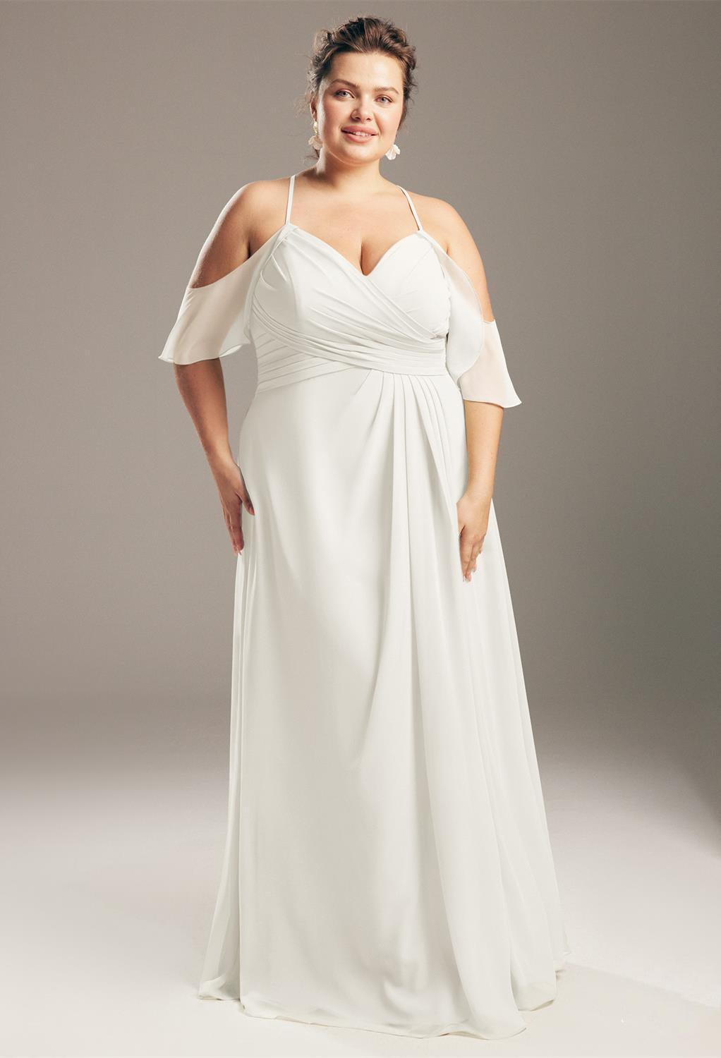 A person is wearing the "Jenifer - Chiffon Bridesmaid Dress - Off the Rack" by Bergamot Bridal, a stunning white gown with an off-the-shoulder design and a ruched bodice, posing against a plain background—a vision that seems straight out of a bridal shop in London, Ontario.