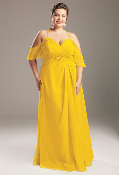 A woman in a vibrant yellow, floor-length chiffon dress with off-shoulder straps smiles gracefully, capturing the sophistication of Bergamot Bridal's affordable selections in London, Ontario. She poses against a simple gray backdrop, echoing the exquisite range of Jenifer - Chiffon Bridesmaid Dresses available off the rack.