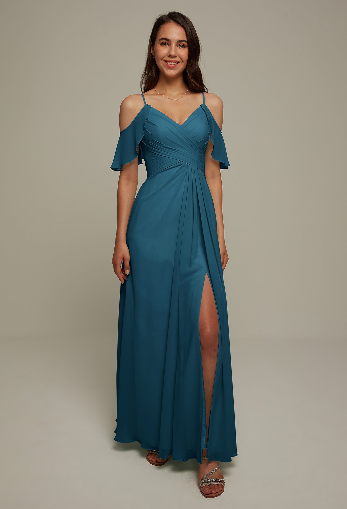 A woman in the Jenifer chiffon bridesmaid dress from Bergamot Bridal, featuring a striking blue hue with an off-shoulder design and a thigh-high slit, stands against a plain background. She's smiling as if she just visited a bridal shop in London, Ontario.