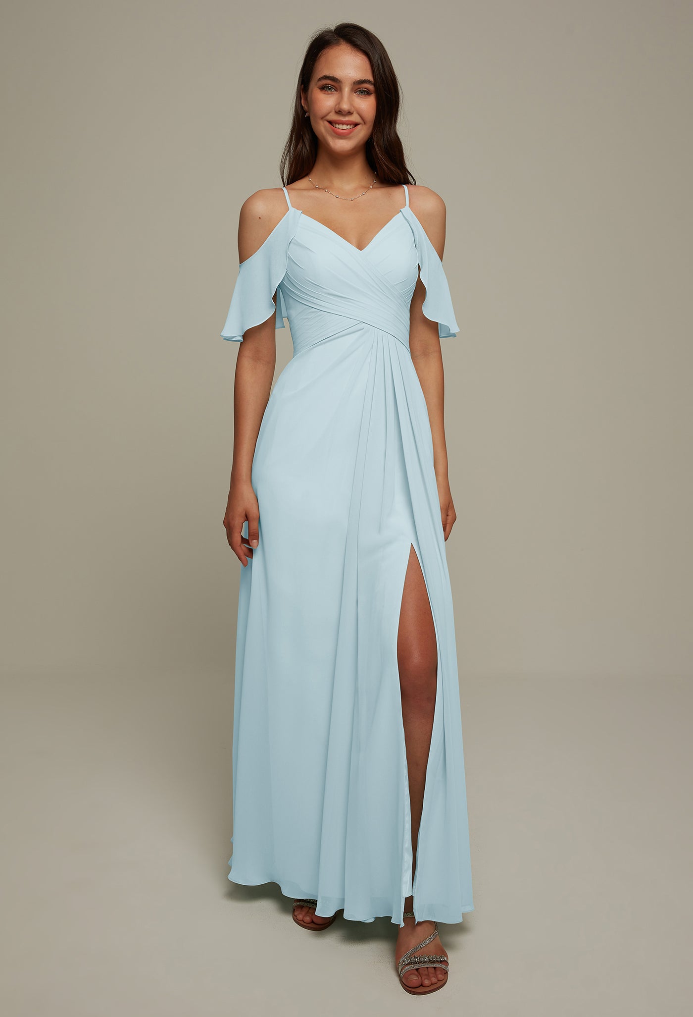 A person is wearing the Jenifer Chiffon Bridesmaid Dress by Bergamot Bridal, featuring a light blue, off-shoulder design with a slit, standing against a plain background reminiscent of bridesmaid dresses found in bridal shops in London, Ontario.