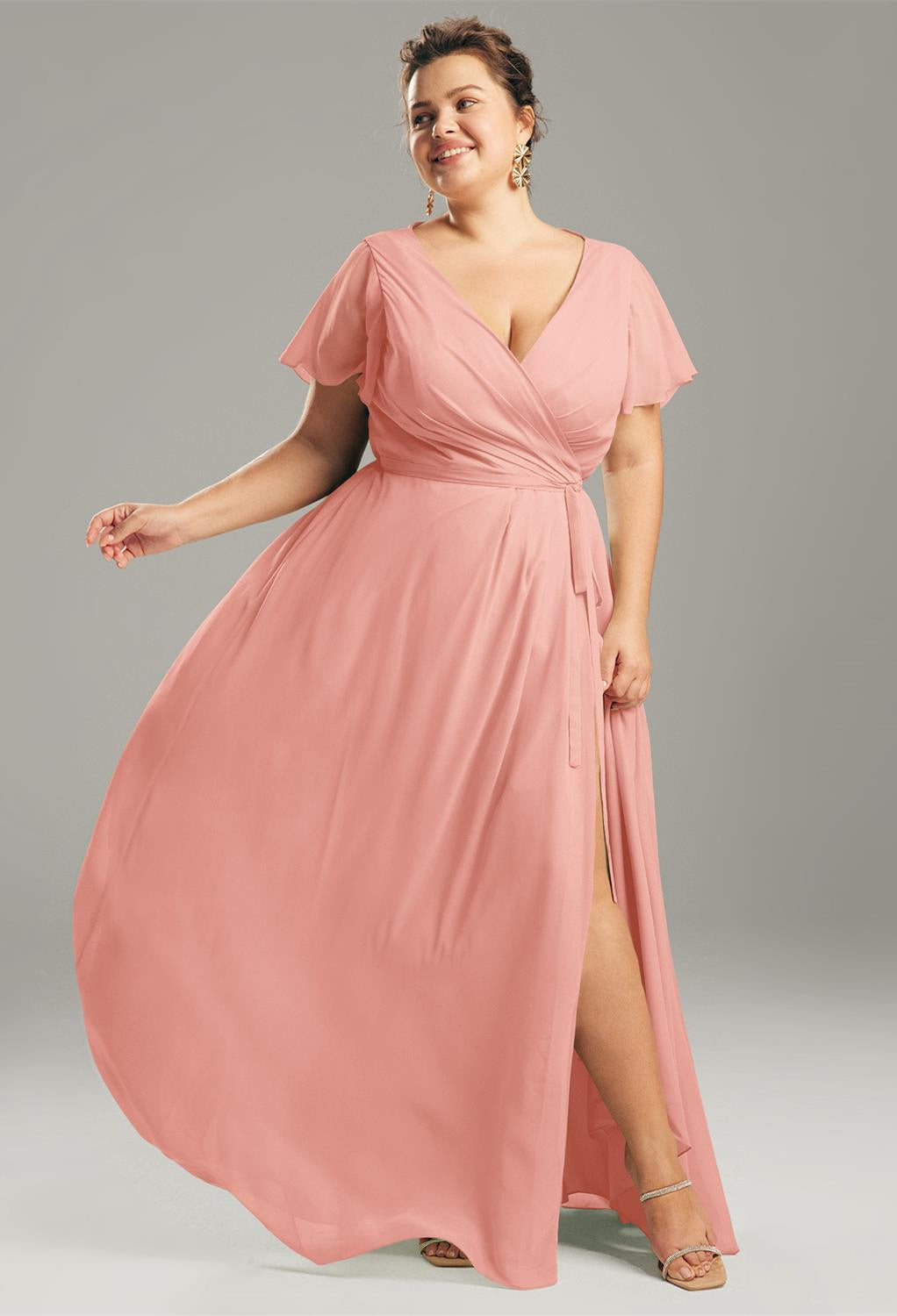 A woman stands confidently, wearing the Ellison - Chiffon Bridesmaid Dress from Bergamot Bridal, featuring a flowing design with a slit. She is accessorized with earrings and sandals, making it the perfect choice for exploring bridal shop options in London, Ontario or effortlessly charming as a bridesmaid.