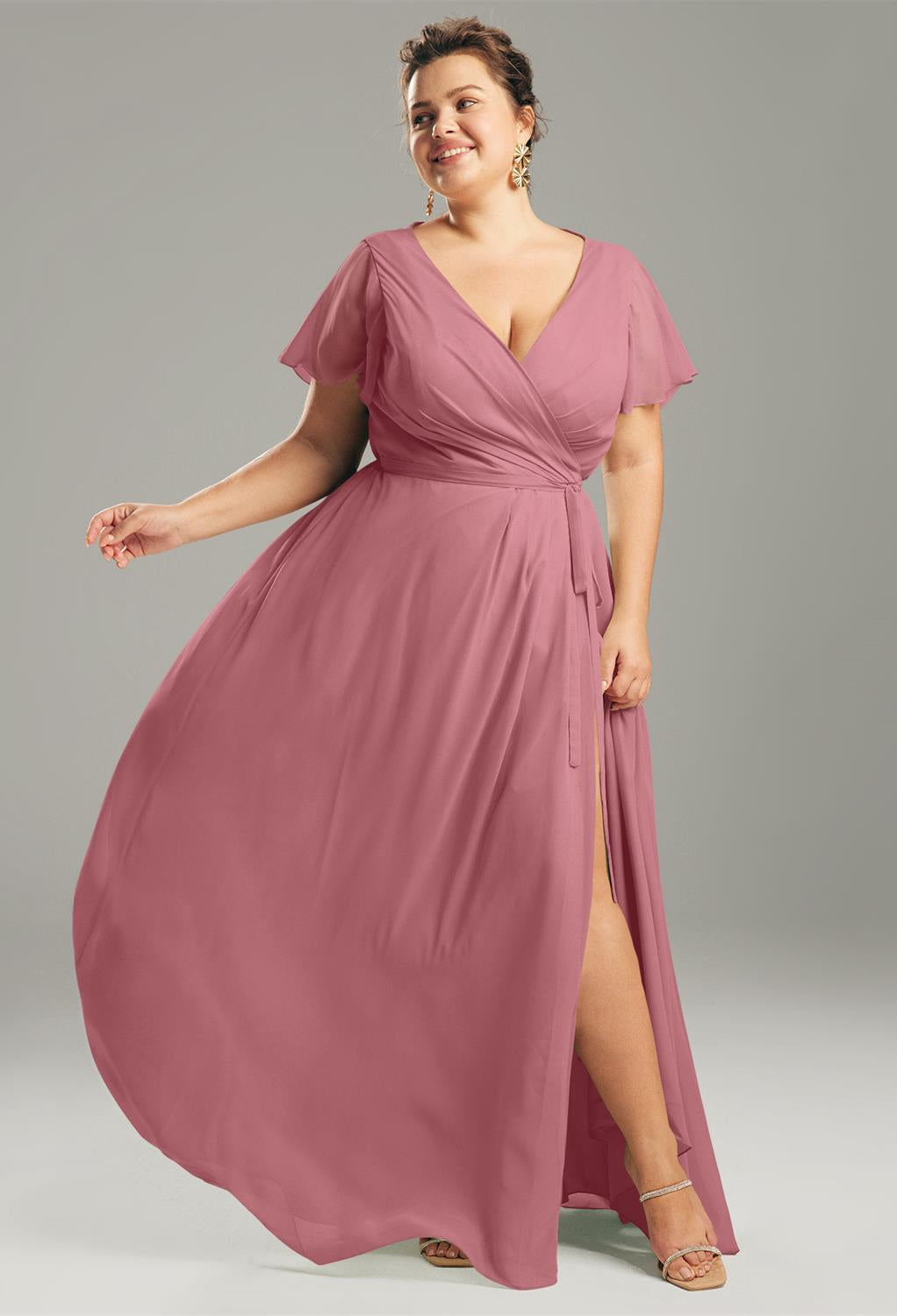 A woman in the Ellison Chiffon Bridesmaid Dress by Bergamot Bridal poses against a gray background in London, Ontario. The pink wrap dress is possibly perfect for bridesmaid occasions, and she pairs it elegantly with heels and earrings.