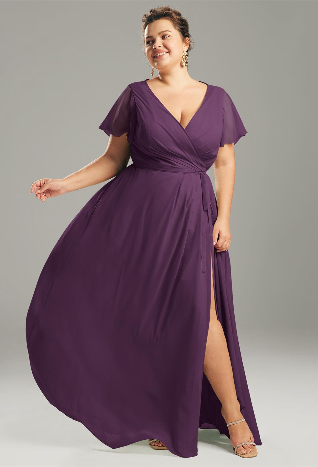 A woman in an airy Ellison chiffon bridesmaid dress from Bergamot Bridal, suitable for any bridal party, poses against a gray background. She accessorizes with elegant earrings and sandals, capturing the sophistication typical of bridal boutiques in London, Ontario.