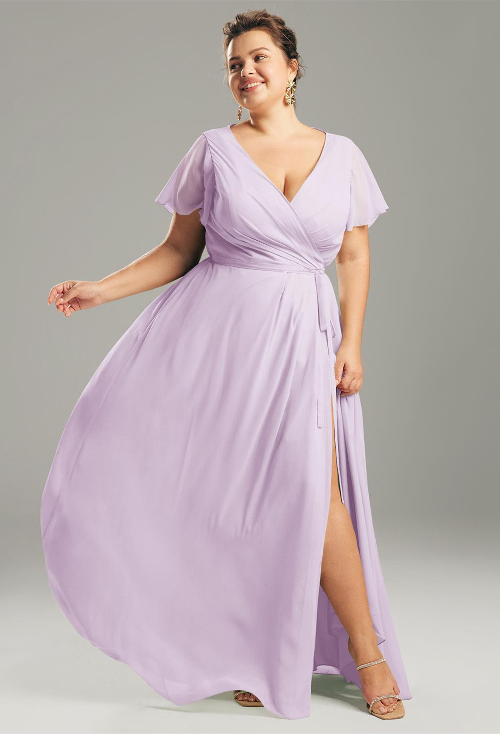 A person wearing the Ellison - Chiffon Bridesmaid Dress from Bergamot Bridal, featuring a flowing lavender design with flutter sleeves, and paired with sandals, stands against a plain background. This dress resembles the elegant bridesmaid dresses often found in bridal shops in London, Ontario.