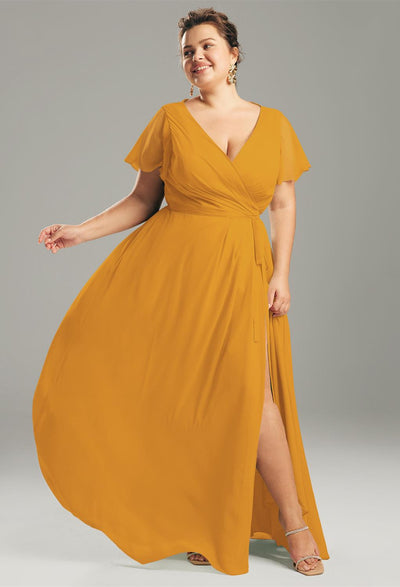 Someone poses against a gray backdrop wearing the Ellison chiffon bridesmaid dress from Bergamot Bridal, showcasing an inspiring choice for stylish bridal shops with its orange hue, flutter sleeves, and elegant slit design.