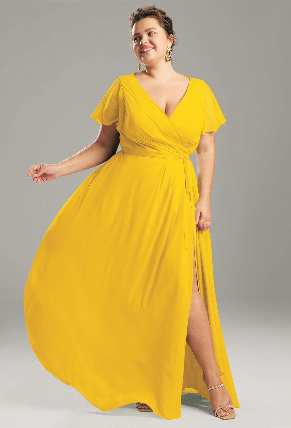 A person smiles while wearing a flowing yellow wrap dress and strappy sandals, resembling the elegant Ellison Chiffon Bridesmaid Dress by Bergamot Bridal, available off the rack in a bridal shop in London, Ontario, as they stand against a plain gray background.