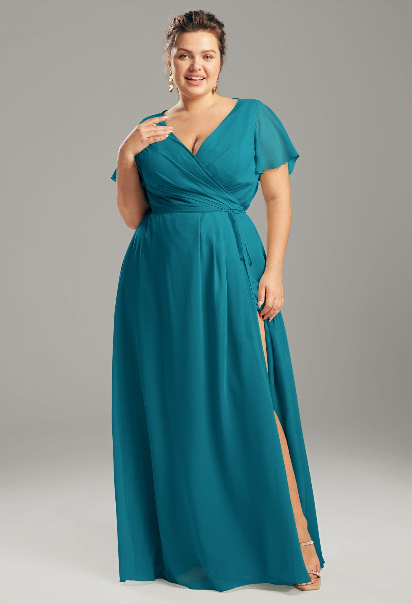 A woman in an Ellison chiffon bridesmaid dress from Bergamot Bridal, featuring a teal hue, flutter sleeves, and a side slit, poses against a gray background, exuding the elegance often seen in London's bridal boutiques.