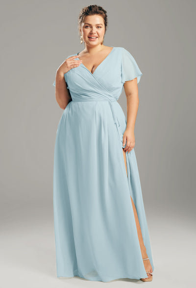 A woman in an Ellison chiffon bridesmaid dress from Bergamot Bridal, featuring a light blue color with short sleeves and a leg slit, smiles against a gray background—a stunning example of the affordable wedding dresses provided by London's top bridal shop in Ontario.