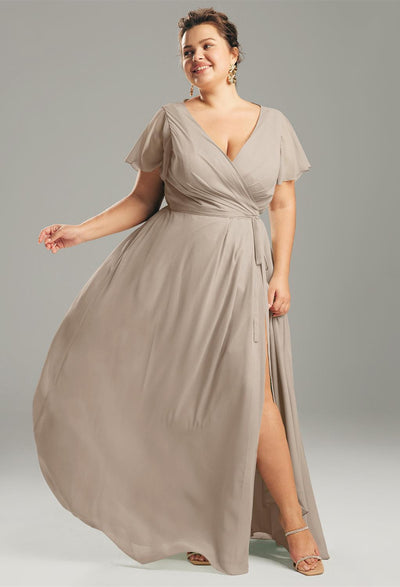 A woman in an Ellison chiffon wrap dress from Bergamot Bridal poses gracefully with one leg visible, reminiscent of the elegant bridesmaid dresses available at bridal shops in London, Ontario. She stands against a gray background, exuding timeless charm.