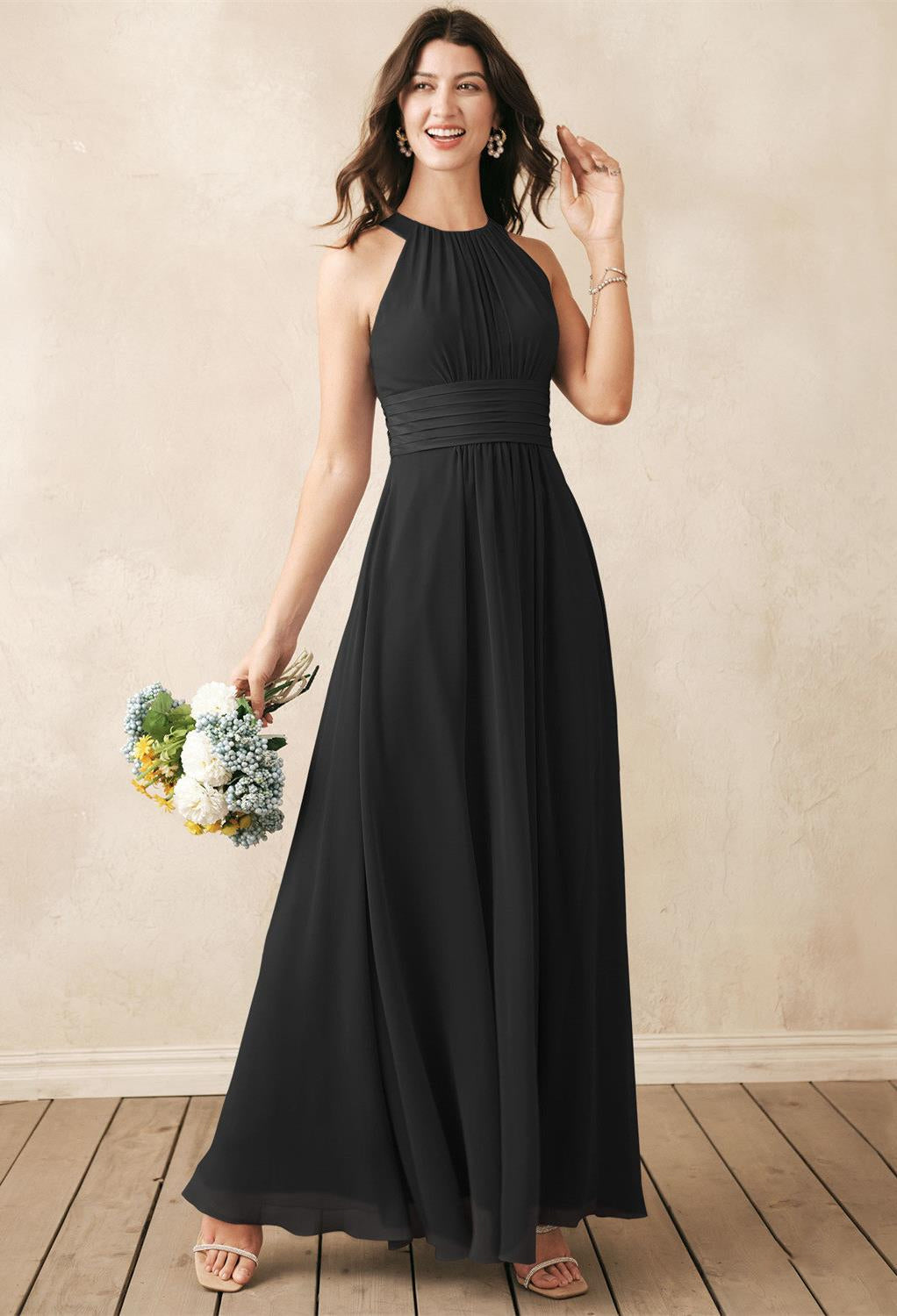 A woman in a flowing Dorian - Chiffon Bridesmaid Dress from Bergamot Bridal stands elegantly on a wooden floor, holding a bouquet of flowers, as if posing amidst the charm of a bridal shop where bridesmaid dresses and affordable wedding gowns are thoughtfully curated.