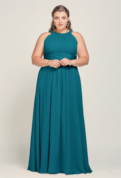 A woman in a teal, floor-length Dorian chiffon gown with a halter neckline from Bergamot Bridal stands elegantly, embodying the glamour of affordable wedding dresses.