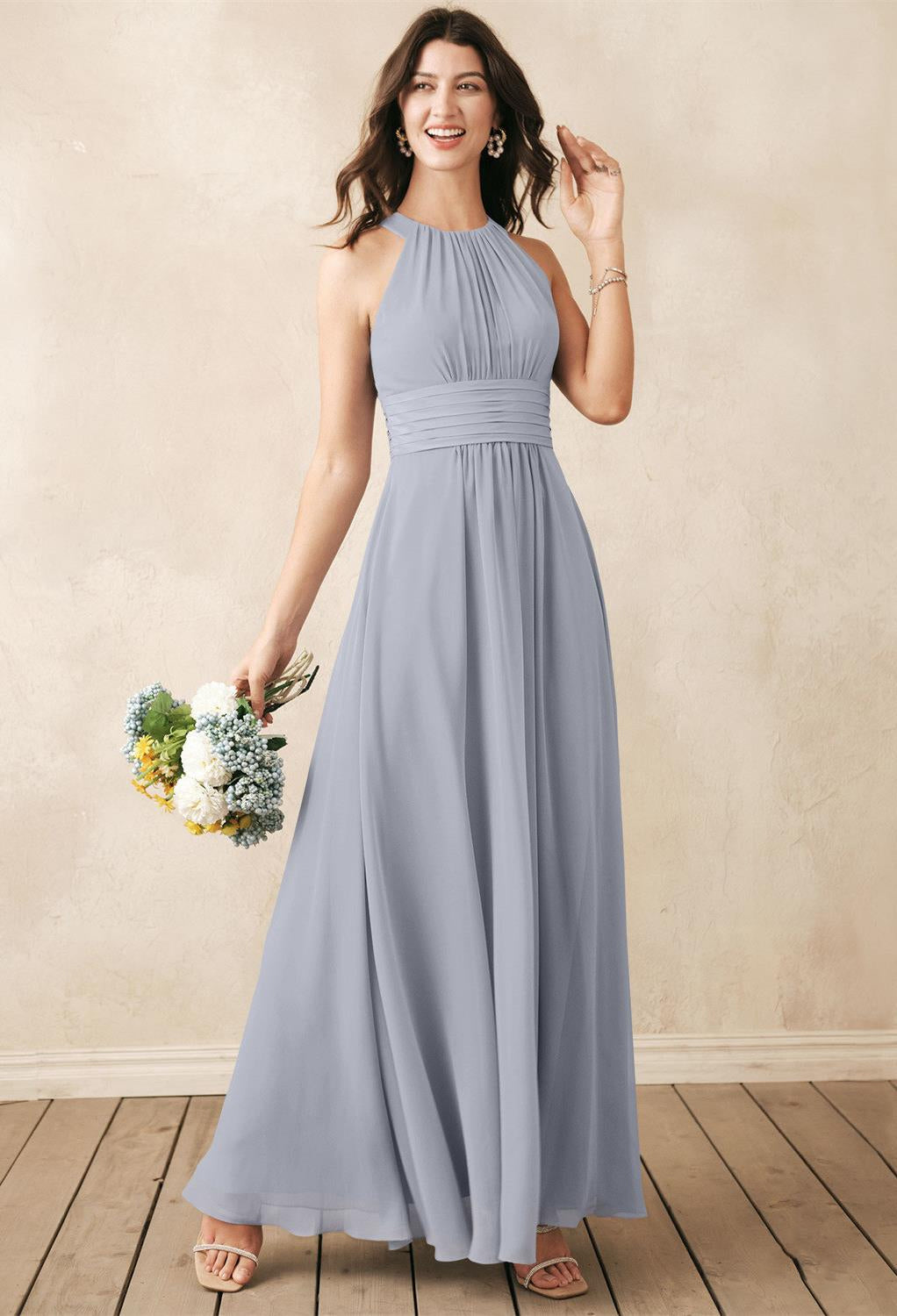 A woman in a long Dorian chiffon bridesmaid dress from Bergamot Bridal in London, Ontario, stands on a wooden floor holding a bouquet of white and yellow flowers, smiling.