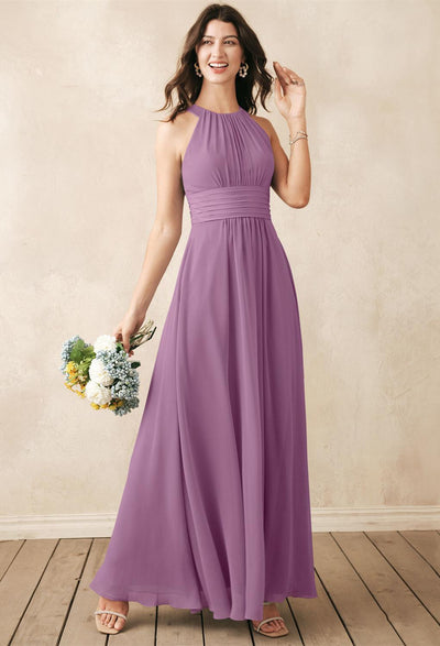 A woman wearing the Dorian - Chiffon Bridesmaid Dress from Bergamot Bridal exudes elegance while holding a bouquet of flowers. Standing on a wooden floor against a textured beige wall, she perfectly exemplifies the grace associated with off-the-rack bridesmaid dresses from the renowned bridal shop in London, Ontario.