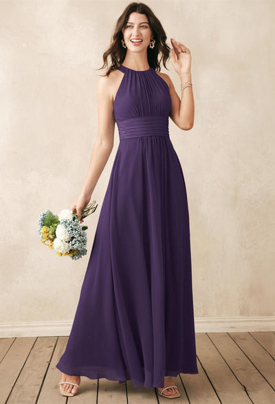 A woman in the Dorian chiffon bridesmaid dress from Bergamot Bridal, a stunning sleeveless purple gown, holds a bouquet of flowers. She stands gracefully on a wooden floor with a textured cream-colored wall serving as the perfect backdrop, evoking the elegance of bridal attire from London, Ontario.