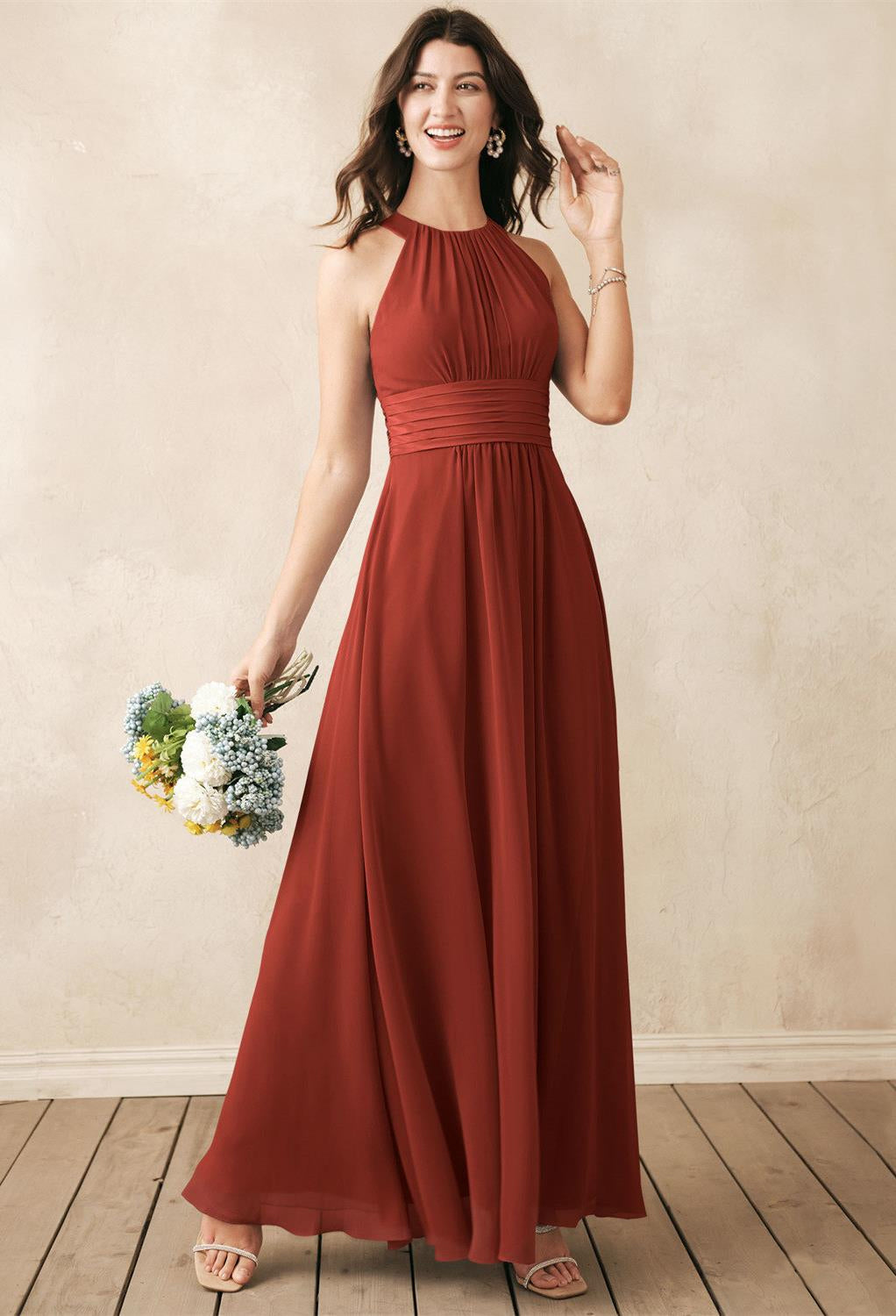 Wearing the Dorian - Chiffon Bridesmaid Dress from Bergamot Bridal, a person in a sleeveless rust hue stands on a wooden floor against a beige wall, holding a small bouquet, showcasing the understated elegance of bridal shops in London, Ontario.