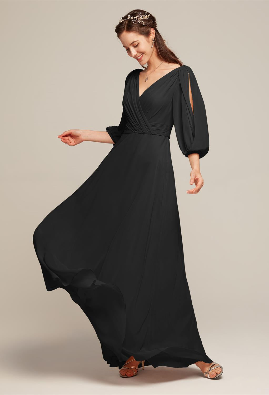 A woman in a long, flowing black dress with a V-neckline stands sideways, smiling slightly. Adorned with a decorative headpiece and wearing sandals, she embodies the elegance of the Polly - Chiffon Bridesmaid Dress by Bergamot Bridal. Ideal for those searching for stylish yet affordable wedding attire in bridal shops around London, Ontario.