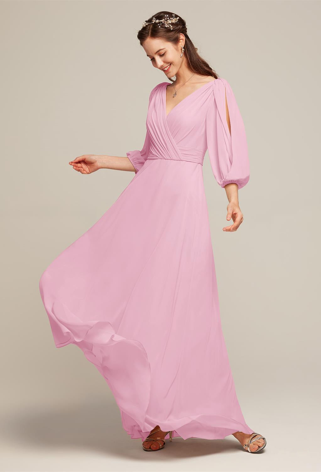 A woman in a Polly chiffon bridesmaid dress from Bergamot Bridal, in a lovely floor-length pink with flowing sleeves, twirls with a smile. Ideal for any bridal shop in London, Ontario, this ensemble truly embodies the spirit of bridesmaid fashion. She finishes her look with a headband and sandals.