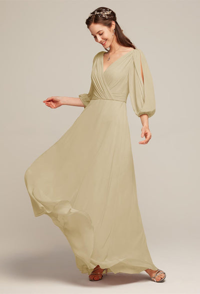 A woman in a long, flowing Polly Chiffon Bridesmaid Dress, featuring puffed sleeves and light beige tones, stands gracefully against a plain background. With sandals on her feet and a slight smile on her face as she holds the dress in her hand, she embodies the elegance found in Bergamot Bridal's affordable off-the-rack collection from London, Ontario.