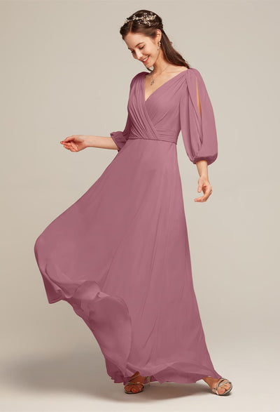 A woman wearing the Polly - Chiffon Bridesmaid Dress by Bergamot Bridal stands against a neutral background, smiling and looking downwards.