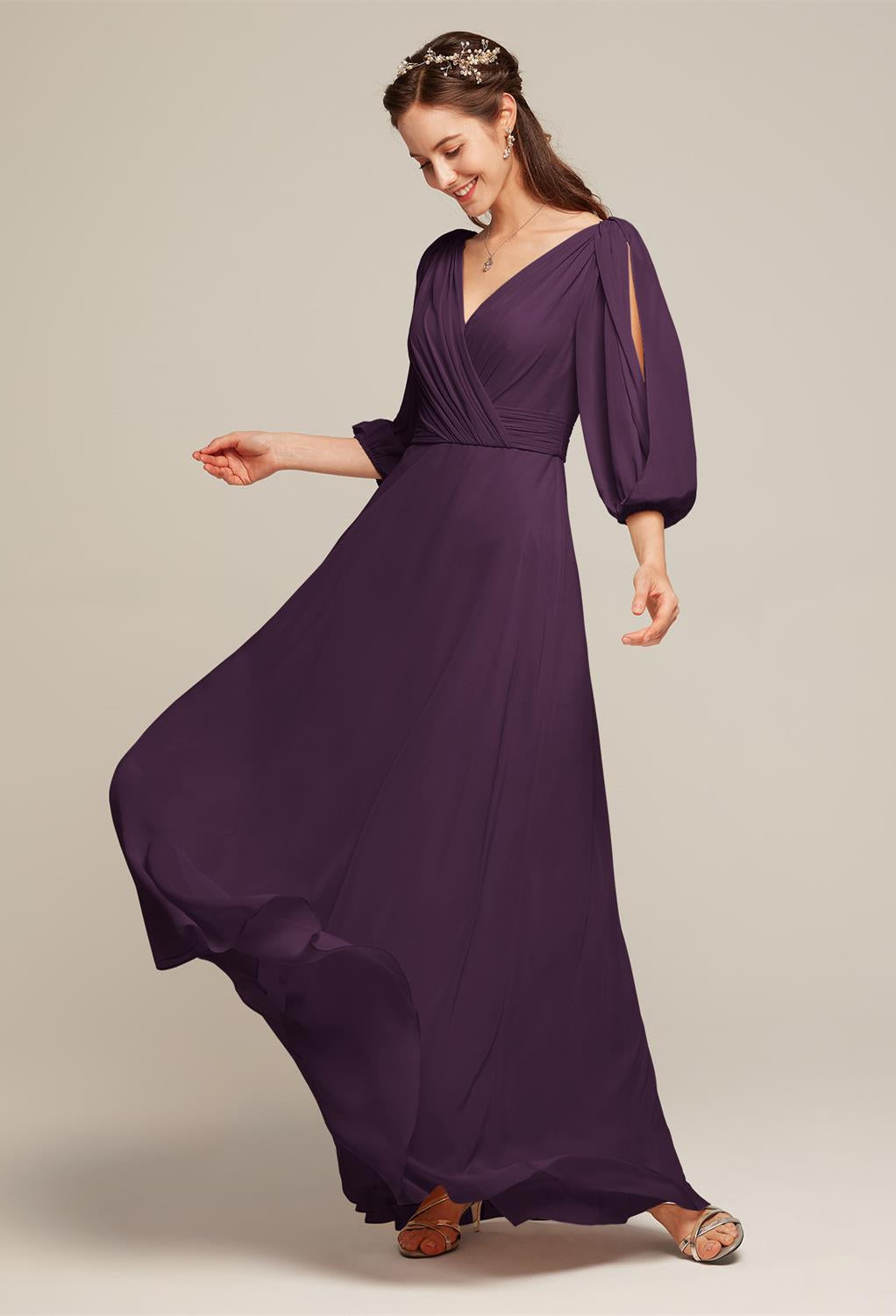 A woman wearing the Polly Chiffon Bridesmaid Dress by Bergamot Bridal, featuring a flowing purple gown with long sleeves and a V-neckline, delicately paired with sandals and jewelry, stands against a simple backdrop, capturing the elegance of off-the-rack bridesmaid dresses from a bridal shop in London, Ontario.