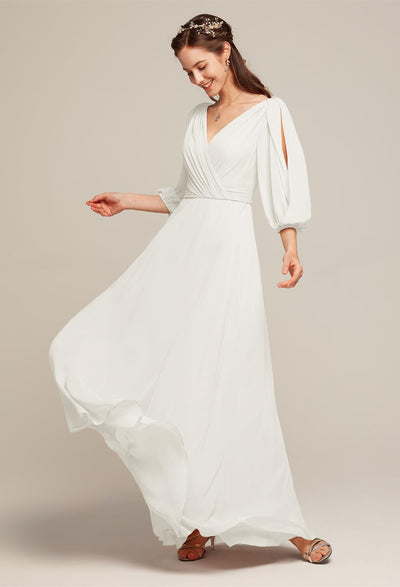 A woman in the sophisticated "Polly - Chiffon Bridesmaid Dress - Off the Rack" by Bergamot Bridal, featuring long, flowing sleeves and a complimentary headpiece, stands gracefully against a light gray backdrop—capturing the essence of a bridal shop's elegance in London, Ontario.