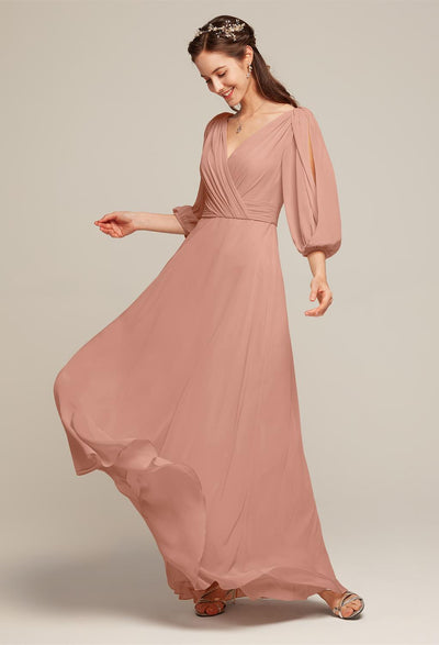 Dressed in the Polly Chiffon Bridesmaid Dress from Bergamot Bridal, a woman elegantly gazes downward while gently holding part of her flowing, peach-colored gown with long sleeves. Her look is completed with a headband and sandals, perfectly capturing the sophistication of this off-the-rack bridesmaid dress against a neutral backdrop.