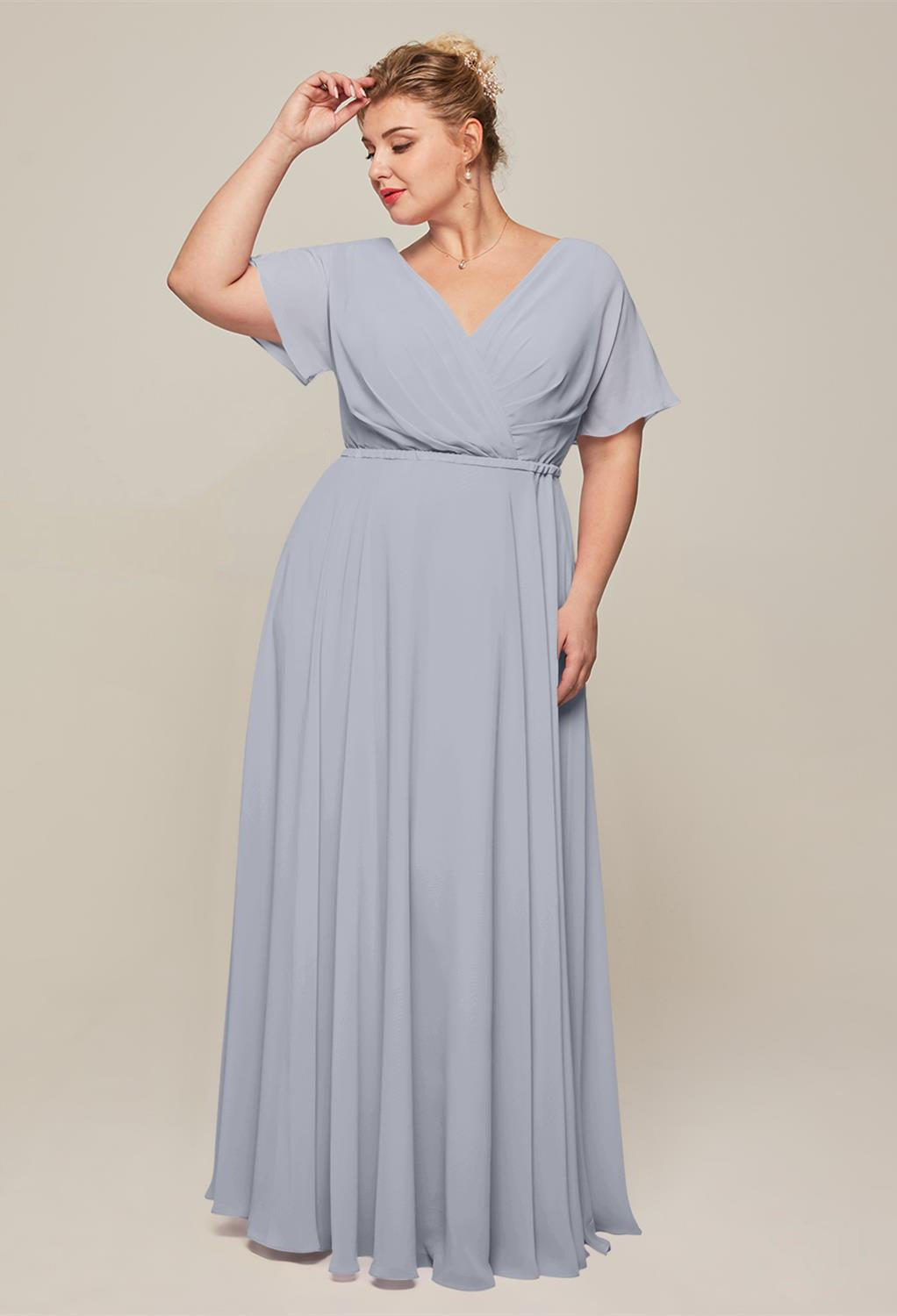 A woman in a light gray floor-length dress poses with her hand touching her hair, capturing the elegance of the Ginny chiffon bridesmaid dress from Bergamot Bridal, a renowned bridal shop in London, Ontario.