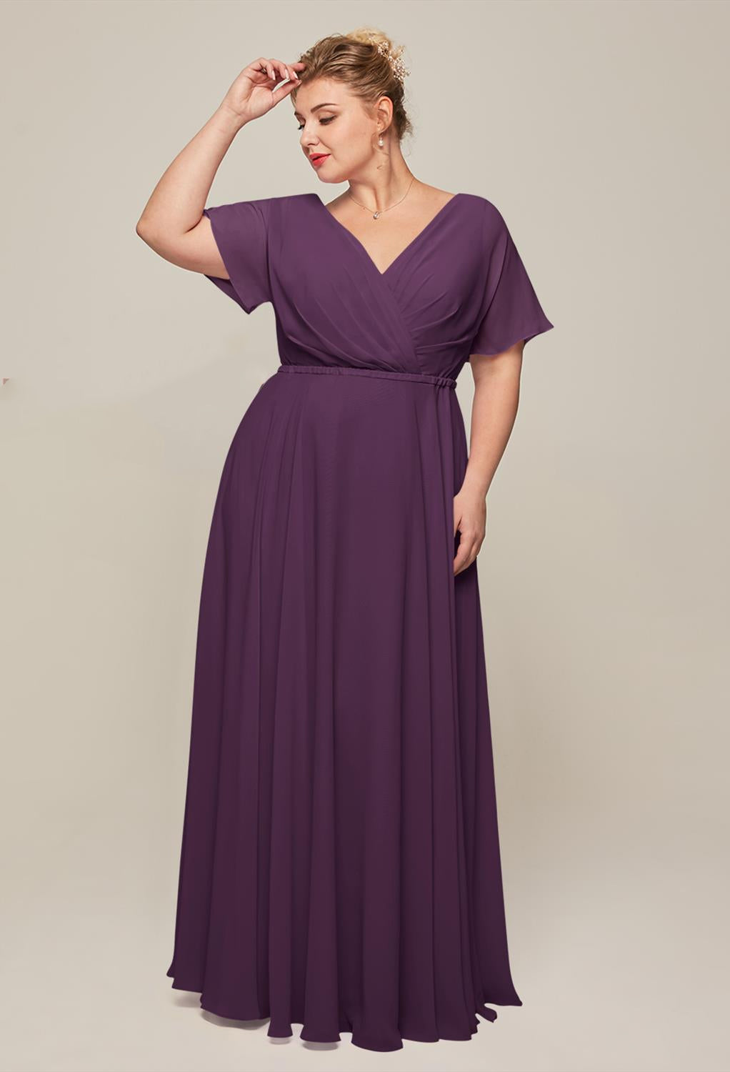 Elegantly channeling the chic styles of Bergamot Bridal's collection, a woman in the Ginny - Chiffon Bridesmaid Dress poses alluringly against a plain backdrop, with one hand gently grazing her hair and the other gracefully resting by her side.