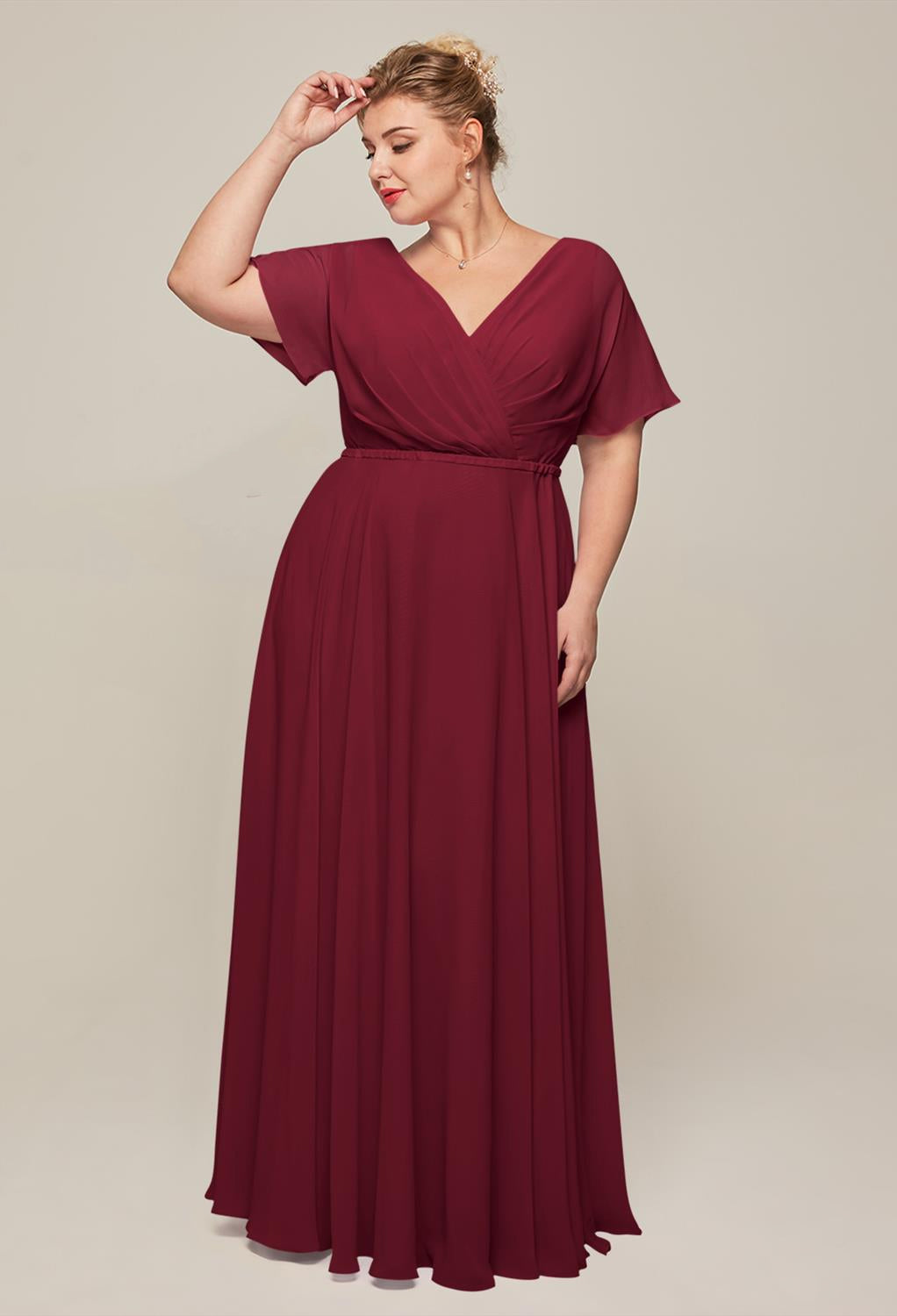 A person stands wearing the Ginny, a chiffon bridesmaid dress by Bergamot Bridal, featuring a floor-length design with short sleeves in burgundy—ideal for a wedding occasion.