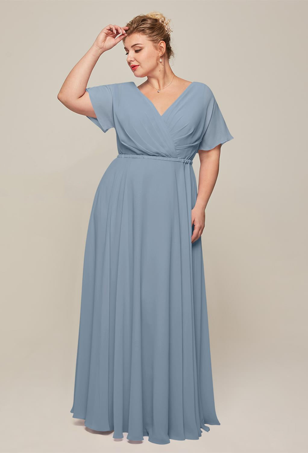 A person wearing the Ginny - Chiffon Bridesmaid Dress by Bergamot Bridal, a light blue, floor-length, short-sleeved gown poses with one hand on their head against a plain background, reminiscent of affordable wedding dresses found at a bridal shop in London, Ontario.