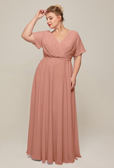 Against a simple backdrop, someone stands wearing the graceful Ginny - Chiffon Bridesmaid Dress by Bergamot Bridal, evoking the elegance of affordable wedding dresses available at a bridal shop in London, Ontario. They glance downward with one hand gently placed in their hair.