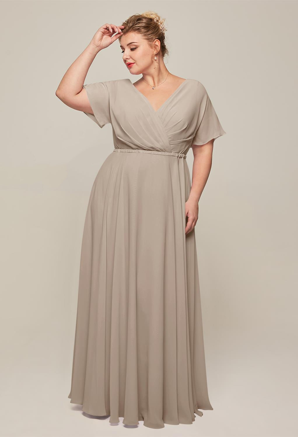 A woman in a light taupe gown, the Ginny Chiffon Bridesmaid Dress by Bergamot Bridal, with a flattering V-neckline and short sleeves stands against a neutral background, gently touching her hair, exuding the same elegance as bridal shop wedding dresses from London, Ontario.