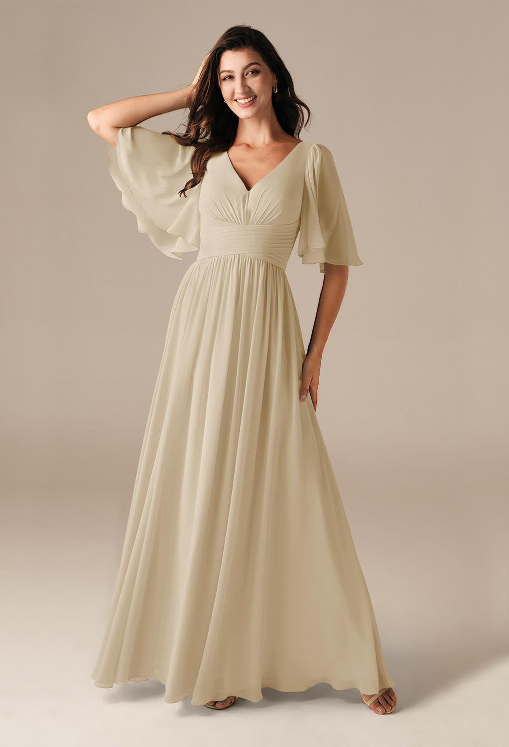 A woman in a long, flowing beige dress featuring short flutter sleeves smiles against a plain background, evoking the elegance of the Kenney Chiffon Bridesmaid Dress by Bergamot Bridal, known for its affordability at their bridal shop in London, Ontario.