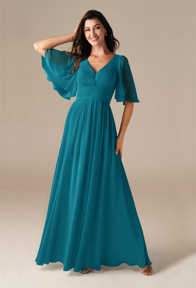 A woman in a teal, floor-length Kenney Chiffon Bridesmaid Dress with flutter sleeves from Bergamot Bridal poses gracefully, her hand on her head. Her elegance captivates like the finest offerings at a bridal shop in London, Ontario.