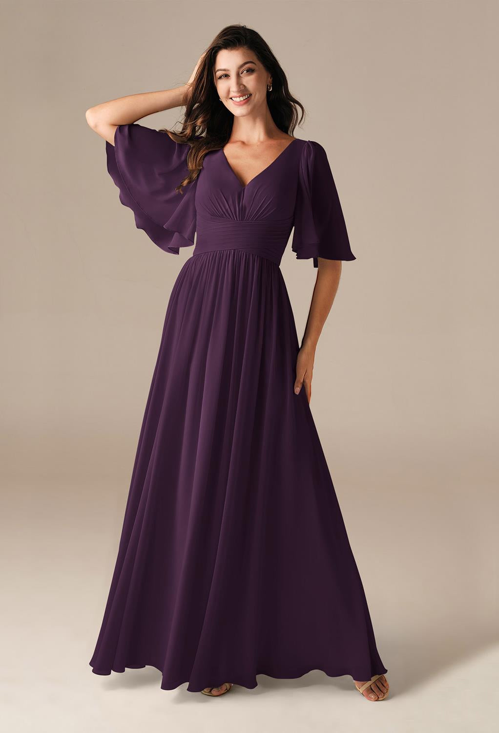 A woman wearing the Kenney chiffon bridesmaid dress by Bergamot Bridal, characterized by its elegant purple hue and flutter sleeves, poses against a plain backdrop, evoking the sophisticated style found in bridal shops throughout London, Ontario.