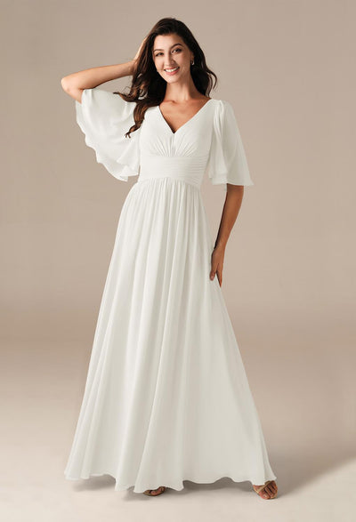 A woman stands gracefully in the Kenney - Chiffon Bridesmaid Dress from Bergamot Bridal, an exquisite off-the-rack gown that echoes the elegant wedding dresses often discovered at a bridal shop in London, Ontario.