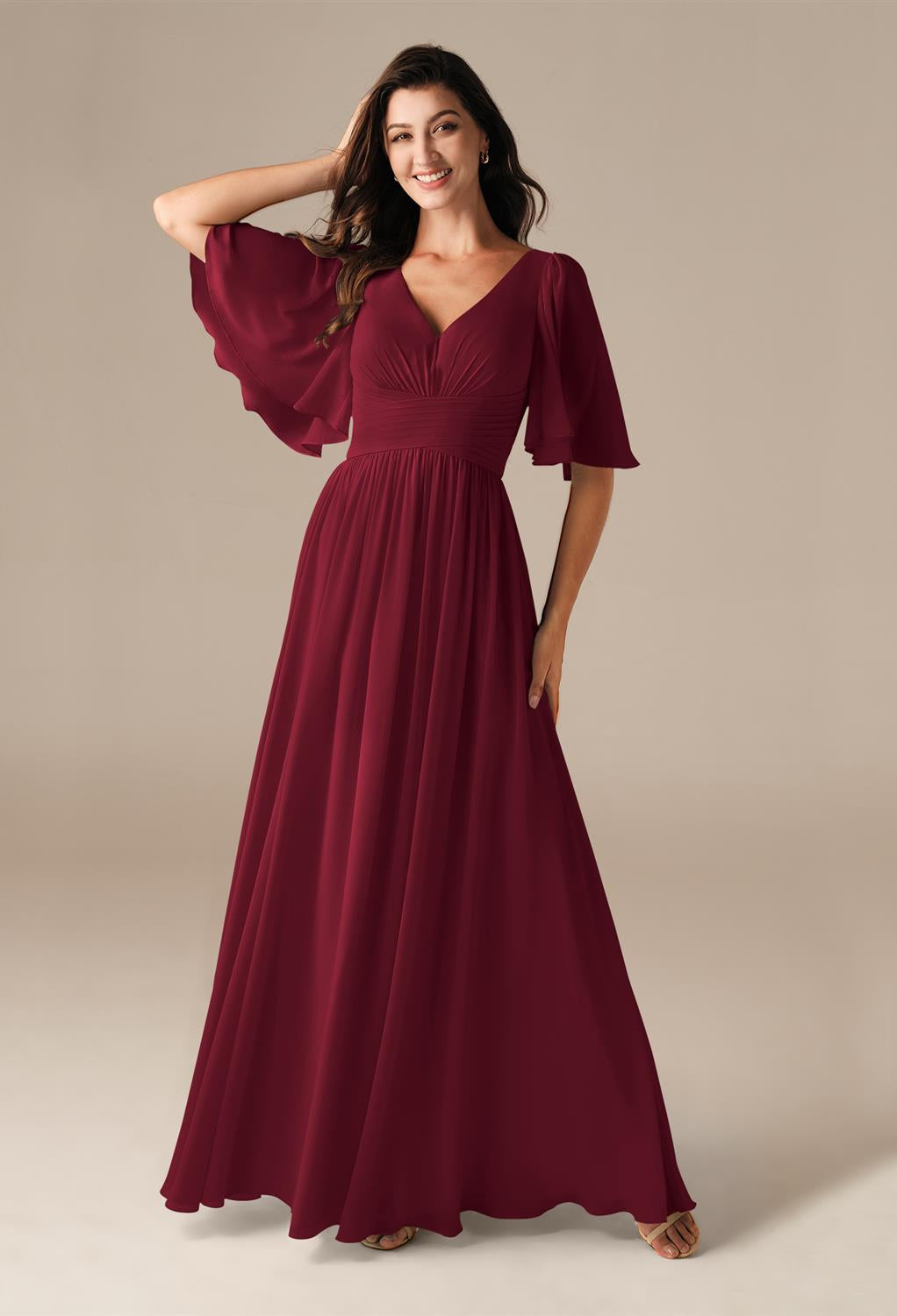 A woman in a flowing burgundy Kenney dress with flared sleeves and a V-neckline stands against a neutral background, reminiscent of the exquisite chiffon bridesmaid dresses from Bergamot Bridal available off the rack in London, Ontario.