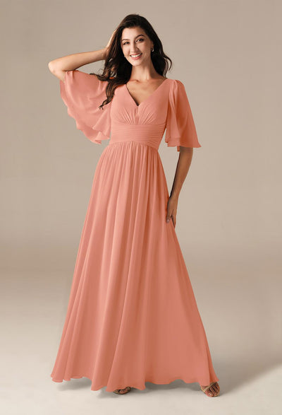 A woman in a flowing gown from Bergamot Bridal, adorned with short flutter sleeves and a delicate peach hue, poses against a neutral background, showcasing the elegance of the Kenney Chiffon Bridesmaid Dress that you can discover at bridal shops in London, Ontario.