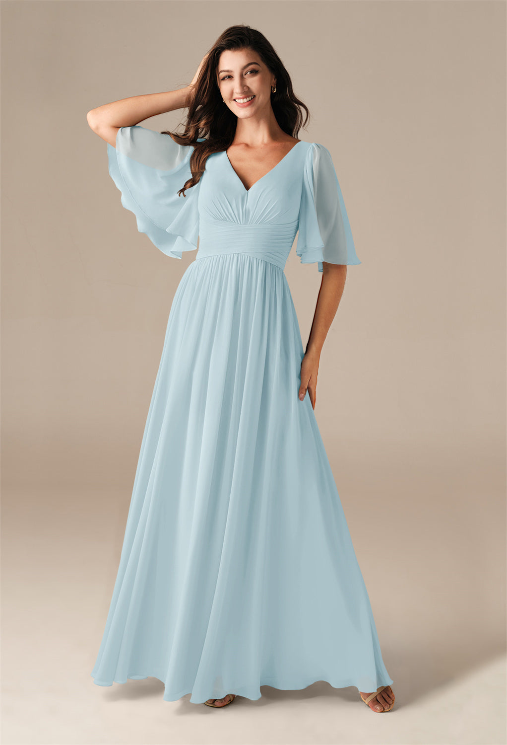 A woman in a light blue gown with flutter sleeves stands gracefully against a neutral background, reminiscent of the elegant Kenney - Chiffon Bridesmaid Dress from Bergamot Bridal, found at the finest bridal shop in London, Ontario.