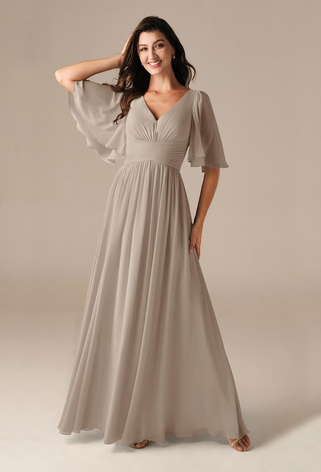 A woman gracefully poses against a neutral backdrop, embodying the charm and sophistication of bridal shops in London, Ontario, wearing the Kenney Chiffon Bridesmaid Dress with flutter sleeves by Bergamot Bridal.