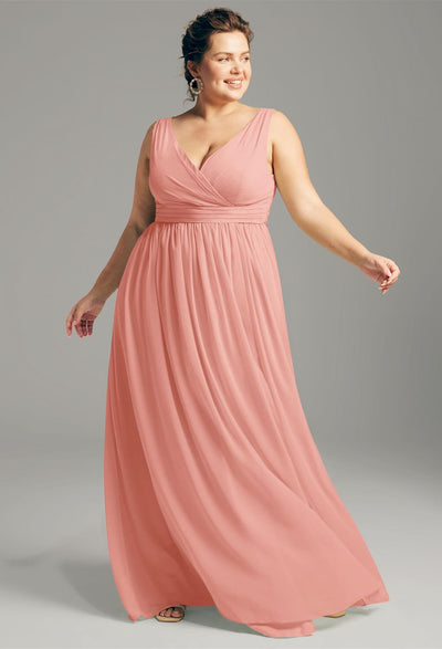 A woman elegantly poses against a gray background, wearing the Hilaria - Chiffon Bridesmaid Dress from Bergamot Bridal. This sleeveless, floor-length pink dress perfectly captures the sophistication typical of bridesmaid dresses available off the rack at this bridal shop in London, Ontario.