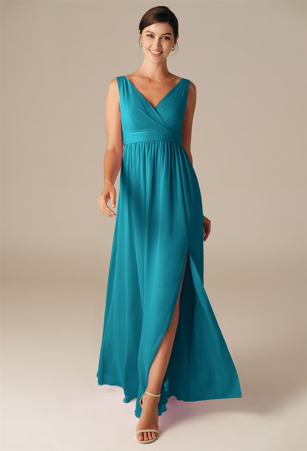 A woman in the Hilaria chiffon bridesmaid dress from Bergamot Bridal, featuring a teal color, sleeveless design, V-neckline, and front slit, stands against a plain background, embodying the elegance typically found at bridal shops in London, Ontario.