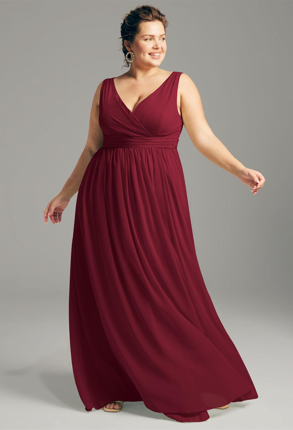 In the refined setting of a bridal shop in London, Ontario, a woman in an off-the-rack Hilaria chiffon bridesmaid dress by Bergamot Bridal stands elegantly against a gray background. Her sleeveless, burgundy floor-length gown complements the sophistication typical of bridesmaid and affordable wedding attire, as she gazes to the side with one hand gently raised.